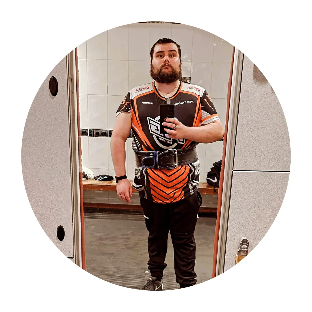 Pictured Callum standing in a locker room, taking a mirror selfie with his phone. He is wearing a black and orange sports jersey, black trousers, and a thick weightlifting belt around his waist.