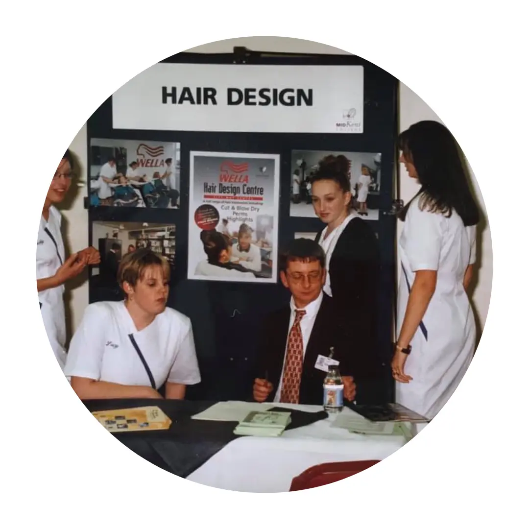 A group of five people gathered around a table at a booth with a sign above that reads 'HAIR DESIGN.' Three women are wearing white uniforms, one woman in black, and one man in a suit and tie is seated at the table looking at some papers.