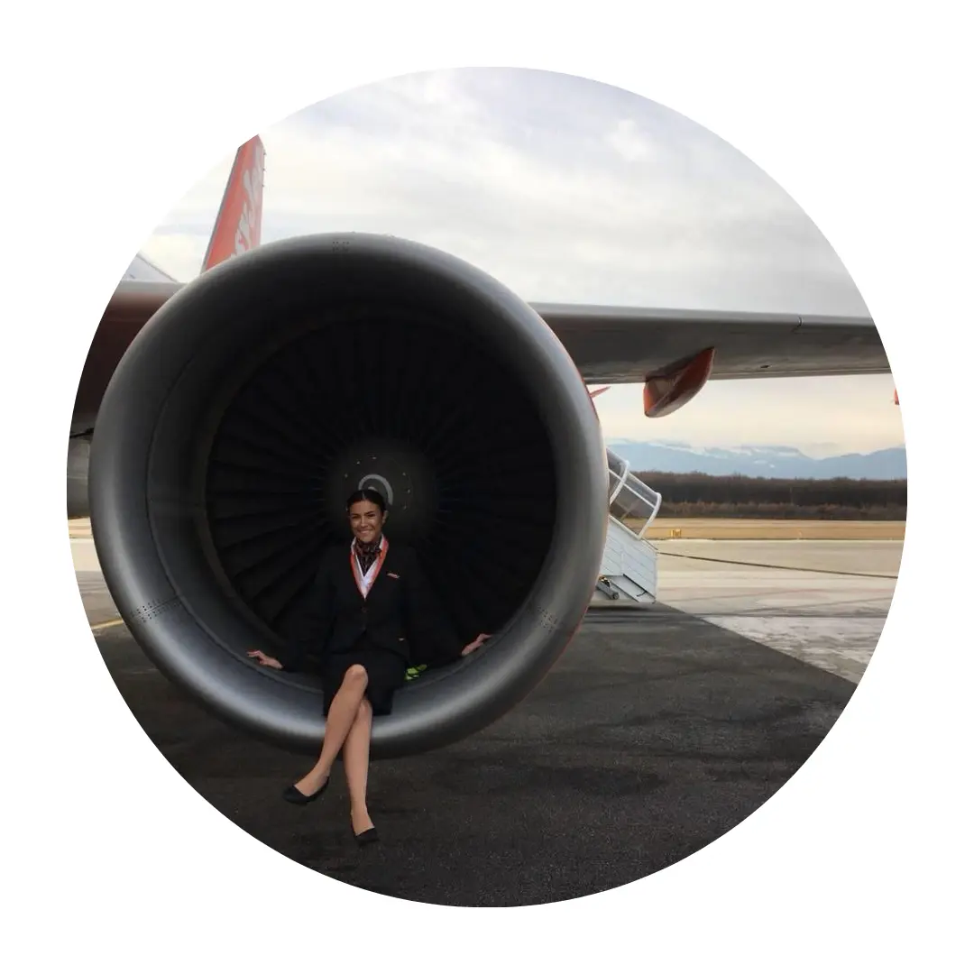 Pictured Paige Collins sitting inside the engine intake of an airplane. She is wearing a flight attendant uniform, and smiling at the camera.