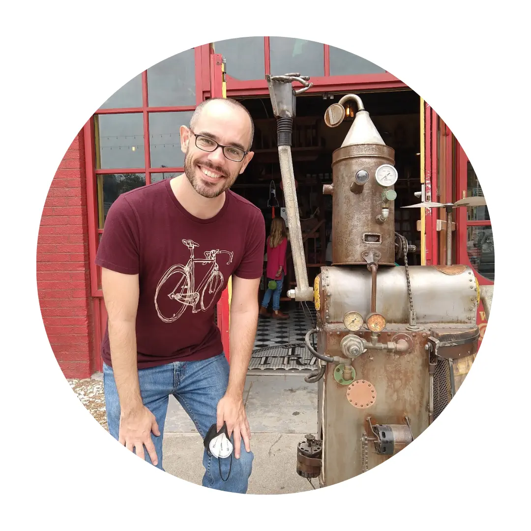 Pictured Sean posing next to a quirky metallic sculpture that resembles a robot. They are outside a building with a red brick wall and red window frames.