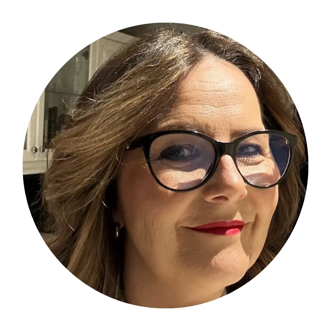 Close-up photo of a smiling woman with light brown hair wearing black-framed glasses and bright red lipstick. She is looking slightly to the side