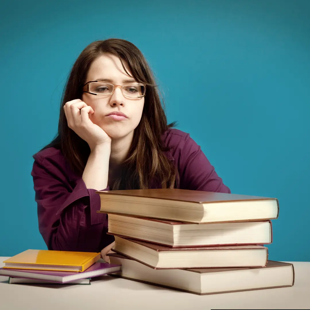 Girl sits staring at a pile of books. Her chin is resting in her hand, and her elbow is on the desk. 