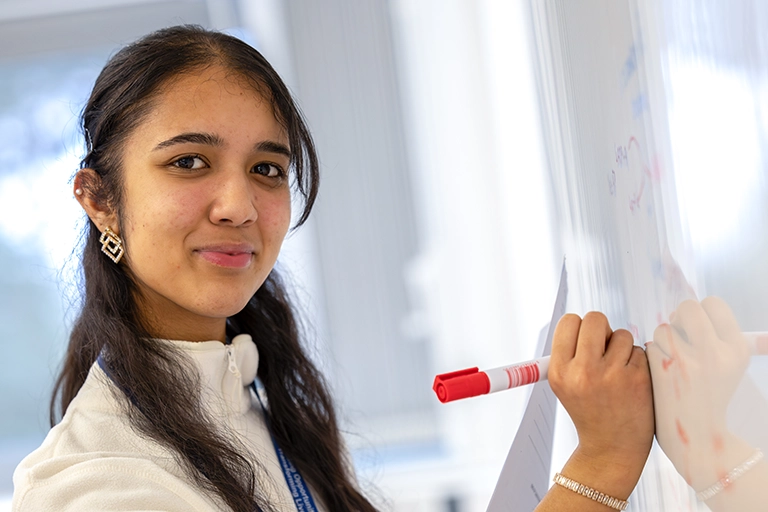 Student writing on the whiteboard whilst smiling at the camera