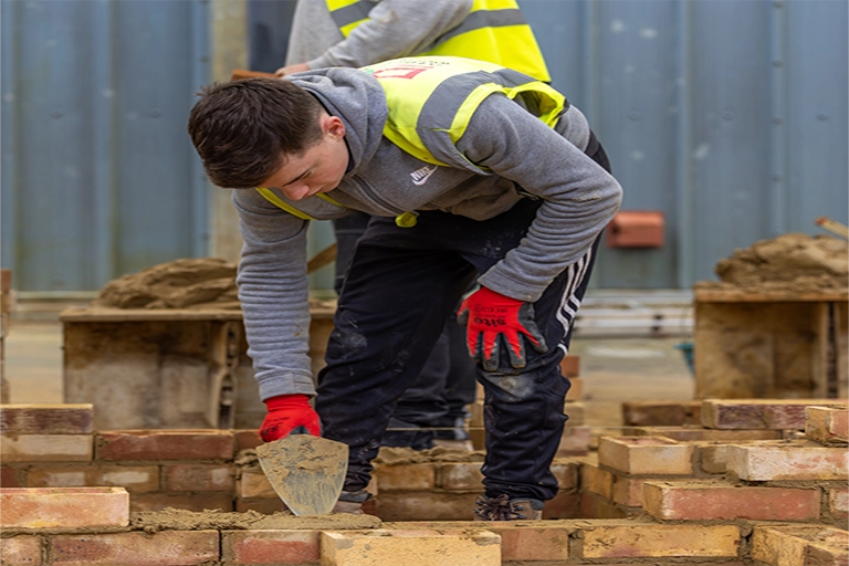 Bricklaying student knelt down in the workshop surrounded by bricks.