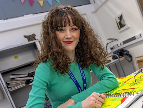 Student wearing a green top and a blue Midkent College lanyard. She has colourful eye makeup with one eye green and the other pink, and she is smiling while sitting at a table with some notebooks and papers in front of her.