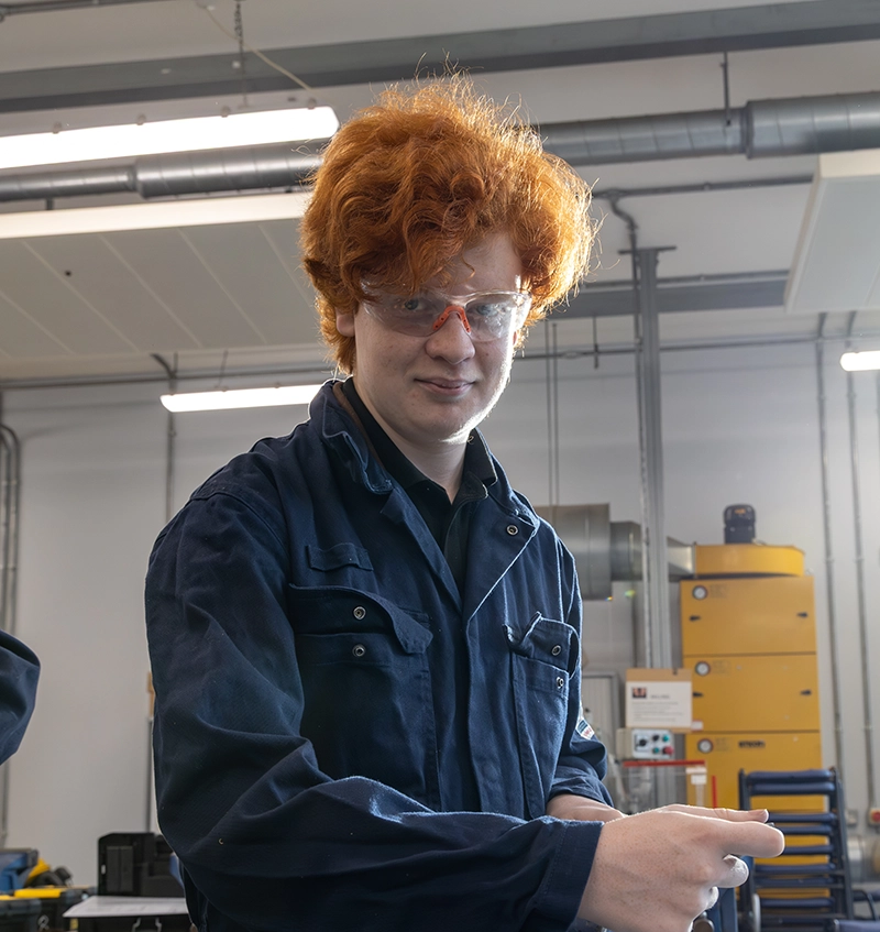 An engineering student wearing safety glasses and blue overalls works in a college engineering workshop, with machinery and equipment visible in the background.