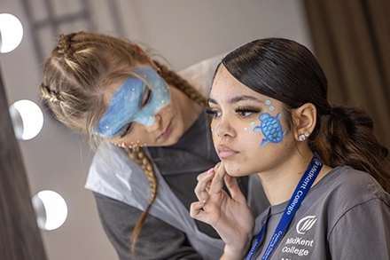 A MidKent College student carefully applies face paint to another student, creating a blue turtle design on her cheek in a makeup studio setting.