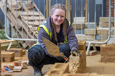 A construction student wearing a high-visibility vest smiles while laying bricks with a trowel at an outdoor building site, surrounded by bricks and scaffolding.
